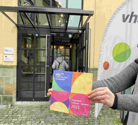 Das Bild zeigt das neue Programmheft, das eine Mitarbeiterin vor unseren Eingang hält - nach dem Motto: Hereinspaziert ins neue Semester.