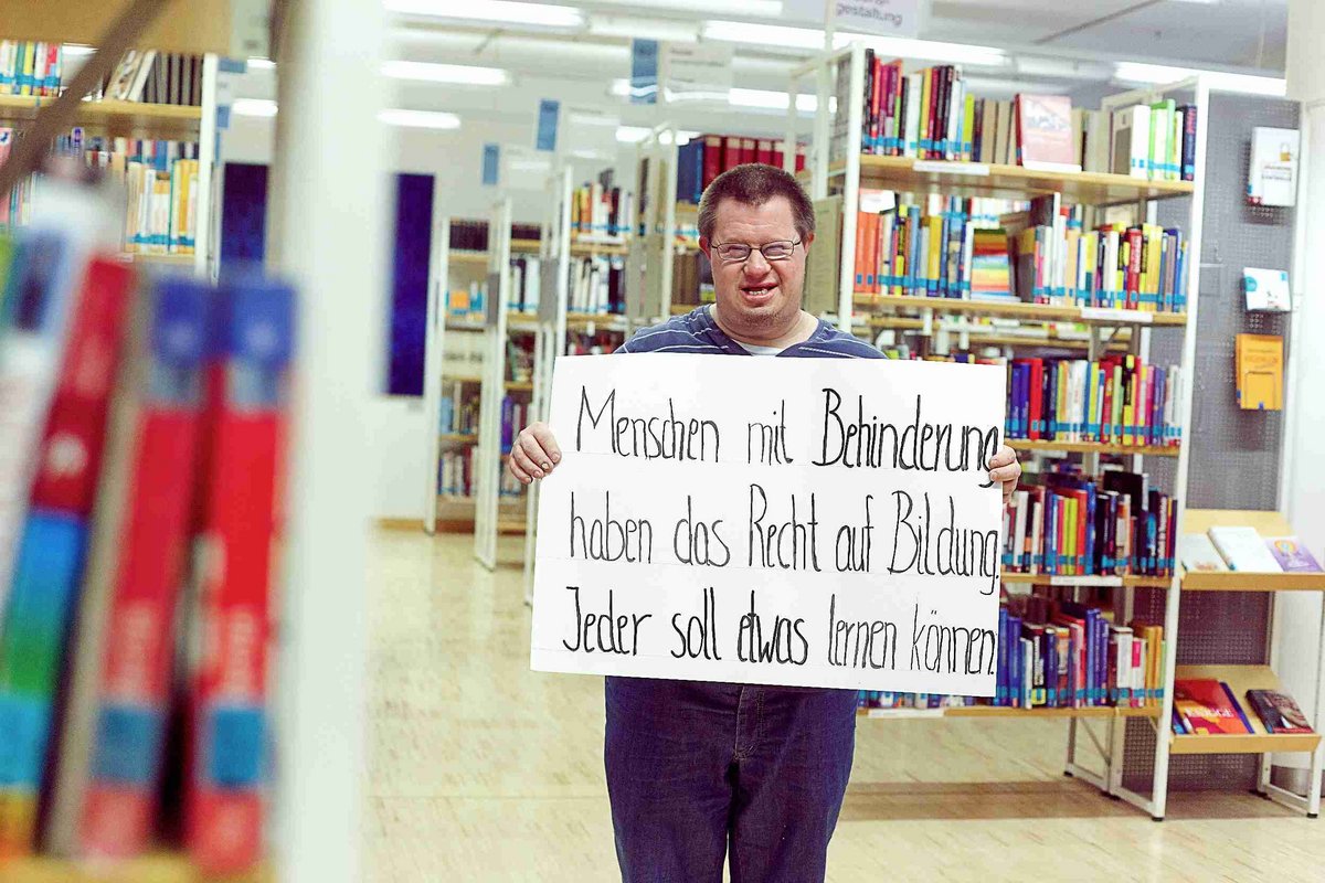 Das Bild zeigt einen jungen Mann mit Down Syndrom in einer Bücherei. Er hält ein Plakat hoch mit dem Text "Menschen mit Behinderung haben ein Recht auf Bildung. Jeder soll etwas lernen können".Alle Menschen haben ein Recht auf Bildung. Foto: Christoph Lilge