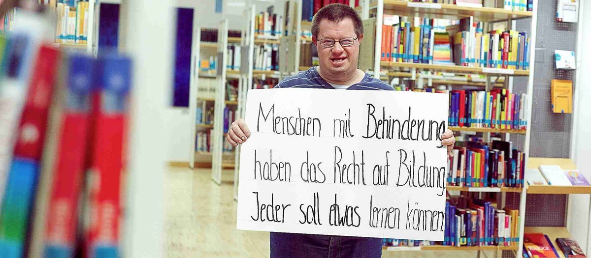 Das Bild zeigt einen jungen Mann mit Down Syndrom in einer Bücherei. Er hält ein Plakat hoch mit dem Text "Menschen mit Behinderung haben ein Recht auf Bildung. Jeder soll etwas lernen können".Alle Menschen haben ein Recht auf Bildung. Foto: Christoph Lilge