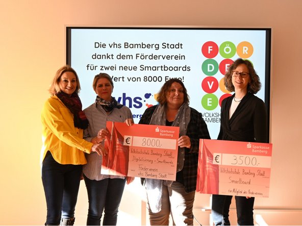Vier Personen halten zwei Spendenschecks in der Hand. Im Hintergrund eines von drei geförderten Smartboards.