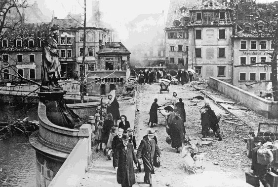 Blick auf die Untere Brücke nach Ende des Zweiten Weltkriegs mit dem Alten Rathaus im Rücken. Zu sehen sind zerstörte Häuser und Menschen, die auf Karren ihre Habseligkeiten transportieren. 